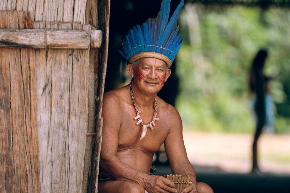 Ancião indígena com cocar de penas azuis tocando flauta sentado na entrada da maloca