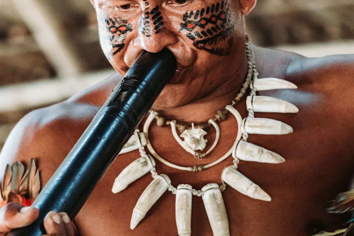 Indígena com cocar azul e pintura facial tocando uma flauta de taboca em close