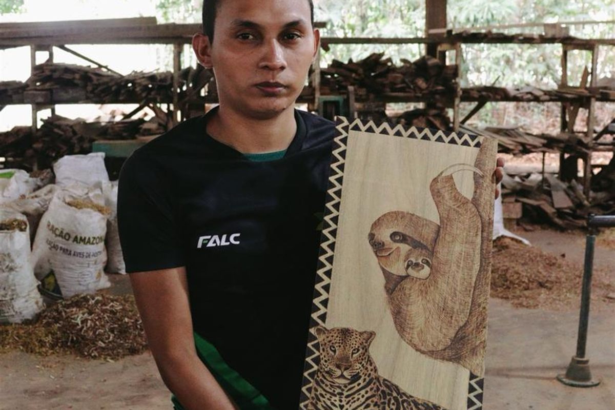 Artesão segurando quadro de pirogravura com onça-pintada e preguiça